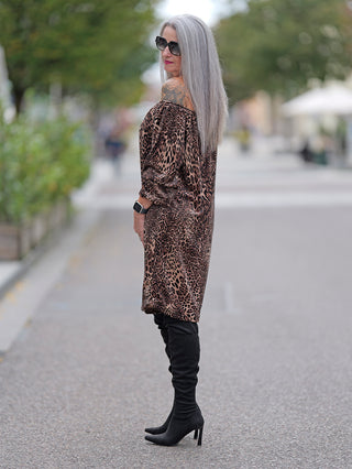 Eine Frau trägt das OFF#DLY Dress Alina leo, ein kurzes Off-Shoulder-Leokleid mit Gummizug am Ausschnitt, kombiniert mit schwarzen Stiefeln auf einem Gehweg.