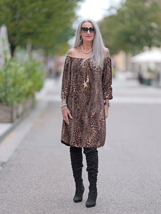 Frau in Sonnenbrille und dem OFF#DLY Dress Alina leo, einem kurzen, schulterfreien Leopardenkleid mit Gummizug am Ausschnitt, kombiniert mit schwarzen Stiefeln und einer Perlenkette.