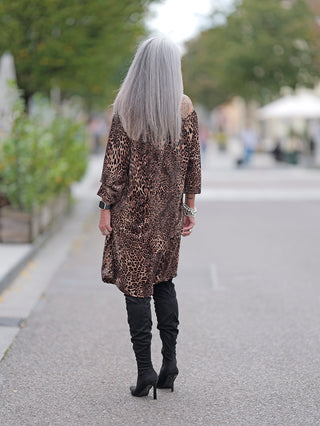 Eine Frau trägt das OFF#DLY Dress Alina leo, ein kurzes, schulterfreies Leopardenkleid mit Gummizug am Ausschnitt, während sie auf einem Gehweg in schwarzen Stiefeln spaziert.