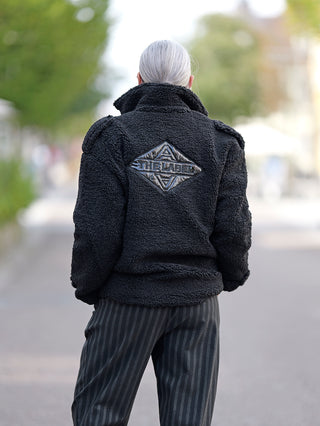 ALIX Teddy Coat in Schwarz, Bikermantel aus weichem Teddystoff mit THE LABEL-Logo auf dem Rücken, Reißverschluss vorn, langen Ärmeln und Schulter-Epauletten.