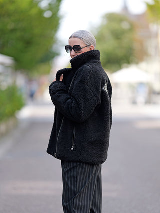 Person trägt den ALIX Teddy Coat in Schwarz, einen Bikermantel aus weichem Teddystoff mit Reißverschluss, langen Ärmeln, Epauletten und Fronttaschen, ideal für stilvolle Straßenmode.
