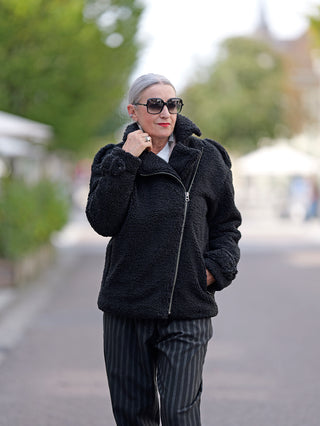 Frau trägt ALIX Teddy Coat schwarz aus weichem Teddystoff mit Reißverschluss und Epauletten, kombiniert mit Sonnenbrille und gestreiften Hosen, für einen modischen Street-Look.