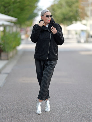 Eine Frau trägt den ALIX Teddy Coat in Schwarz, einen Bikermantel aus weichem Teddystoff mit Reißverschluss und Epauletten, kombiniert mit Sonnenbrille und gestreiften Hosen.
