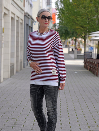 Frau auf dem Gehweg trägt das OFF#DLY Ringelshirt Nena burgundy/white, ein schmales Longsleeve mit seitlichem Zipper, kombiniert mit Jeans und Sonnenbrille für einen lässigen Street-Fashion-Look.