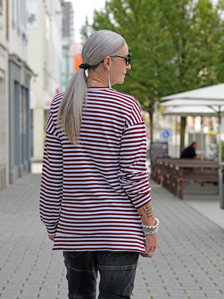 Frau im OFF#DLY Ringelshirt Nena burgundy/white, schmal geschnittenes Longsleeve mit seitlichem Zipper, trägt eine gestreifte Bluse im Freien, kombiniert mit lässiger Street-Fashion.