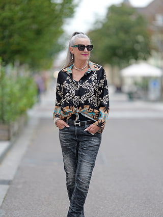 Frau trägt OFF#DLY Bluse Rosella mit Printmix und Knopfleiste, kombiniert mit Jeans, während sie auf einem Bürgersteig geht. Sichtbare Accessoires: Sonnenbrille und Halskette. Bluse ist locker geschnitten.