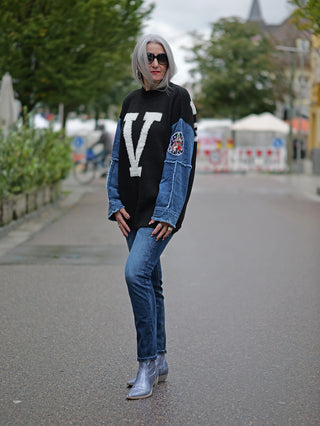 Eine Frau trägt die TAKE 2 Chiba Denim slim Jeans mit schmaler Passform und Midblue Waschung auf der Straße. Kombiniert mit silbernen Stiefeln und einem schwarzen Pullover mit weißem V.