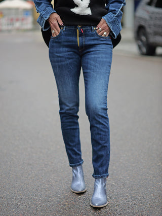 Frau trägt TAKE 2 Chiba Denim Slim Jeans. Schmale Passform mit 5 Taschen in Midblue Waschung. Kombiniert mit silbernen Schuhen. Modelgröße: 172 cm, trägt Größe 28.