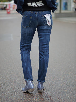 Person trägt TAKE 2 Chiba Denim Slim Jeans mit Etikett in der Gesäßtasche, kombiniert mit Stiefeln. Die Jeans hat eine schmale Passform und ist in einer Midblue Waschung gehalten.