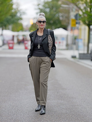 Eine Frau trägt die OFF#DLY Pant Clara khaki mit Gummibund und seitlichen Einschubtaschen, während sie auf der Straße läuft. Sie kombiniert die Hose mit einem schwarzen Jacke und modischen Accessoires.