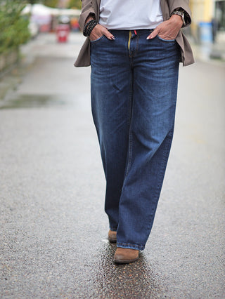 Person trägt TAKE 2 Sendai Denim Wide Leg Jeans mit weitem Schnitt und 5-Pocket-Design. Sichtbar sind die Beine in blauen Jeans und braunen Schuhen, Hände in den Taschen.