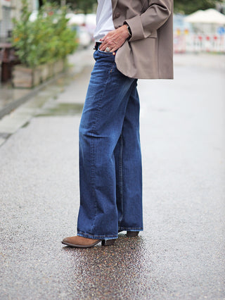 Person trägt TAKE 2 Sendai Denim Wide Leg Jeans; weite Passform, 5-Pocket-Design, Midblue Waschung, kombiniert mit Jacke und Stiefeln. Ideal für einen lässigen Street-Fashion-Look.