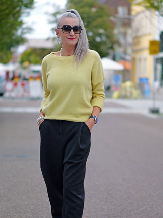Eine reife Frau trägt den OFF#DLY Pullover Nina yellow, einen weichen, leicht kastigen Pullover mit 3/4-Ärmeln, kombiniert mit lockeren schwarzen Hosen, während sie in einer urbanen Umgebung steht.