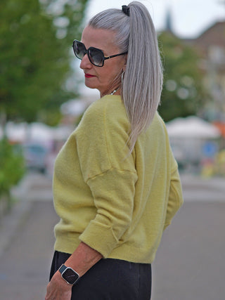 Eine Frau trägt den OFF#DLY Pullover Nina yellow, einen weichen Rundhalspullover mit 3/4 Ärmeln, während sie draußen steht. Der Pullover hat einen lässigen, modernen Look mit hochgeschobenen Ärmeln.