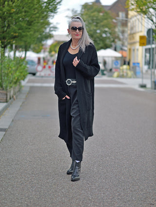 Eine Frau trägt die OFF#DLY Clara Strickjacke lang schwarz, kombiniert mit Sonnenbrillen und Stiefeln, während sie auf der Straße geht. Die Jacke hat eine grobe Strickstruktur und seitliche Taschen.