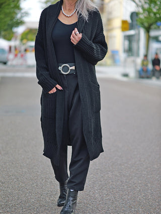Eine Frau trägt die OFF#DLY Clara Strickjacke lang black, eine lange Grobstrickjacke mit seitlichen Taschen, kombiniert mit schwarzen Hosen und Stiefeln.