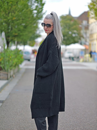 Eine Frau trägt die OFF#DLY Clara Strickjacke lang black, eine lange Grobstrickjacke mit seitlichen Taschen, während sie draußen eine Sonnenbrille und einen schwarzen Mantel präsentiert.