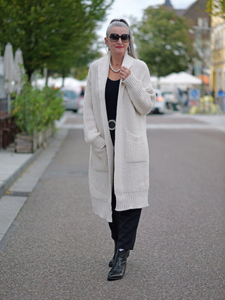 Eine Frau trägt die OFF#DLY Clara Strickjacke lang white, kombiniert mit einer Sonnenbrille, während sie auf einem Gehweg spaziert. Die Jacke hat seitliche Taschen und ist grob gestrickt.