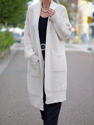 Frau trägt die OFF#DLY Clara Strickjacke lang white, eine lange grobgestrickte Jacke mit seitlichen Taschen, kombiniert mit einem schwarzen Kleid.