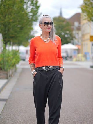 Eine Frau trägt den OFF#DLY Kurzpulli Renata orange mit V-Ausschnitt auf dem Gehweg, kombiniert mit schwarzer Hose und Sonnenbrille. Der Pullover ist schmal und kurz geschnitten.