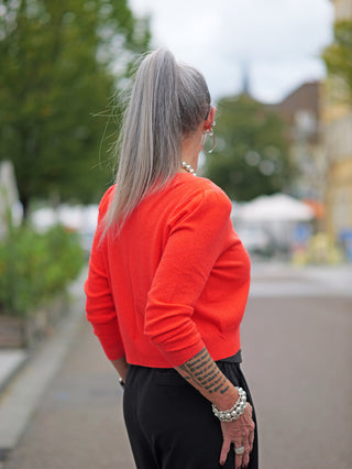 Eine Frau trägt den OFF#DLY Kurzpulli Renata orange, einen schmalen, kurzen Pullover mit kleinem V-Ausschnitt, sichtbar im Outdoor-Setting. Der Pulli ist tailliert und stilvoll.