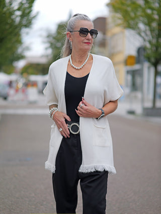 Frau trägt die OFF#DLY Strickjacke Sabrina creme mit Fransen, ohne Knopfleiste, kombiniert mit schwarzem Kleid und Sonnenbrille, zeigt modischen Straßenstil.