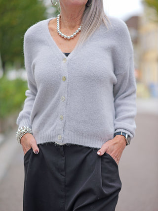 Frau trägt OFF#DLY Weste Fabiola grey, eine kurze Strickjacke mit V-Ausschnitt und Knopfleiste, kombiniert mit schwarzer Hose. Im Fokus: Strickmuster und Passform.