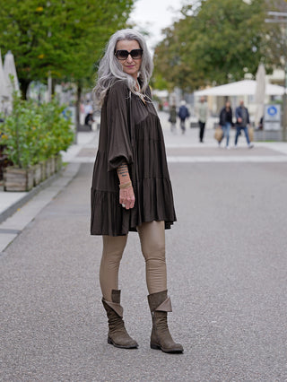 Eine Frau trägt die OFF#DLY Fake Leder Legging Frida in Taupe, kombiniert mit einem braunen Kleid. Sie hat lange graue Haare und trägt Sonnenbrillen, passende Mode für die Straße.