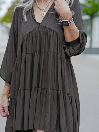 Eine Frau trägt das OFF#DLY Kleid Karin, ein kurzes A-Linien-Kleid mit V-Ausschnitt, während sie ein Handy hält und eine herzförmige Halskette sowie eine Armbanduhr trägt.