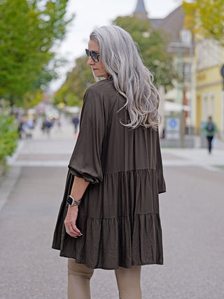 Frau trägt das OFF#DLY Dress Karin, ein kurzes A-Form Kleid mit V-Ausschnitt. Sie hat lange graue Haare und Sonnenbrille, aufgenommen in einem modischen Straßenumfeld.
