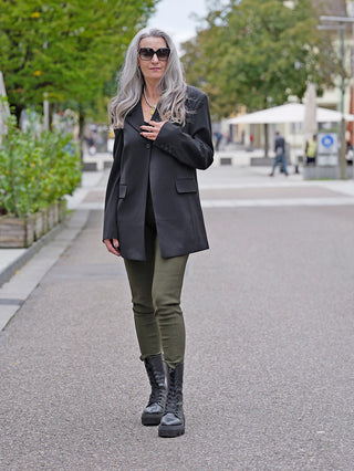 Frau auf Gehweg trägt OFF#DLY Legging Sonja khaki, kombiniert mit schwarzem Mantel und Stiefeln. Leggings aus Viskose-Baumwolle, mit seitlichen Taschen und elastischem Tragekomfort.