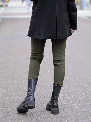 Eine Person trägt die OFF#DLY Legging Sonja khaki mit seitlichen Einschubtaschen, kombiniert mit schwarzen Stiefeln und einem schwarzen Mantel.