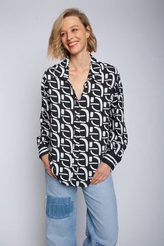 Emily Oversized Bluse in schwarz/weiß, getragen von einer lächelnden Frau. Die Bluse hat überschnittene Schultern, eine Knopfblende, geometrischen Print und eine Manschette mit Streifenbordüre.