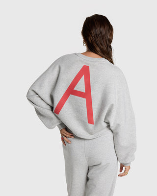 Frau trägt den ALIX Badwing A Sweater grey/melange mit rotem A auf dem Rücken. Kombiniert mit passender Jogginghose für bequemen Look.