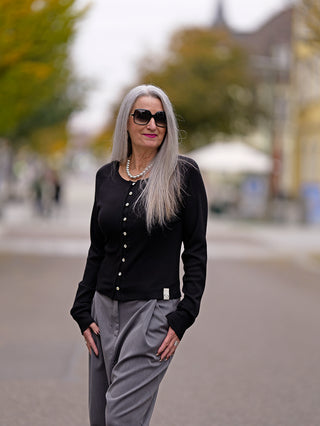 Frau mit grauen Haaren trägt Cotton Candy Ladies Cardigan Lea Schwarz mit auffälliger Knopfleiste und Perlenkette, kombiniert mit Sonnenbrille.