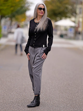 Frau auf der Straße trägt den Cotton Candy Ladies Cardigan Lea in Schwarz, mit auffälliger Knopfleiste, kombiniert mit grauen Hosen und schwarzen Stiefeln; Sonnenbrille als Accessoire.