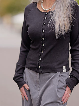 Frau trägt den Cotton Candy Ladies Cardigan Lea in Schwarz mit auffälliger Knopfleiste, kombiniert mit einer Halskette. Hände sind in den Taschen. Modell ist 172 cm groß, trägt Größe S.