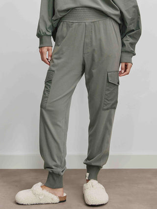 Eine Person trägt die Cotton Candy Ladies Pant Lorna olive mit seitlichen Taschen und elastischem Bund, kombiniert mit einem flauschigen weißen Hausschuh.