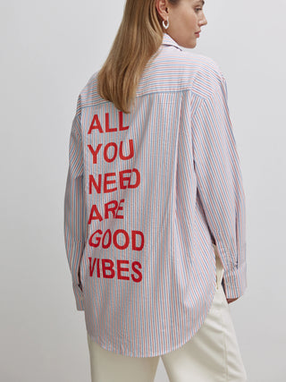 Frau trägt die Cotton Candy Bluse Zoe multicolor, gestreift mit rotem Rückenprint ALL YOU NEED ARE GOOD VIBES. Lässiger Look mit Knopfleiste, perfekt für den Alltag.