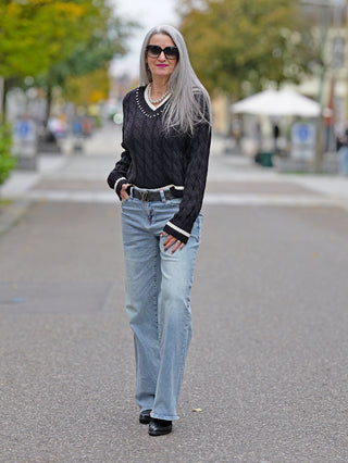 Eine Frau trägt den OFF#DLY Pulli Berta black mit V-Ausschnitt und Zopfmuster, verziert mit Strassteinen, kombiniert mit Jeans und schwarzen Schuhen auf dem Gehweg.