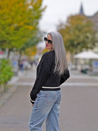 Eine Frau trägt den OFF#DLY Pulli Berta black mit Zopfmuster, V-Ausschnitt und Strasssteinen, kombiniert mit Jeans und Sonnenbrille, während sie im Freien steht.