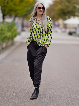 Frau in Emily Shirtbluse green sand mit kleinem Kragen und V-Ausschnittblende, lässig auf dem Bürgersteig gehend, betont durch Lurex Tape und Retro-Print, passend zu ihrer stilvollen Straßenmode.
