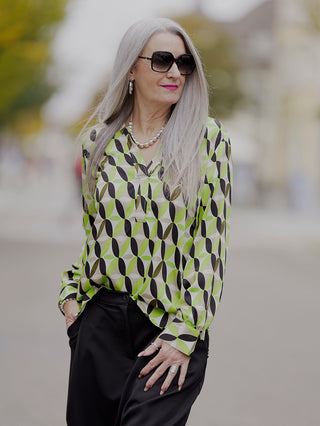 Emily Shirtbluse green sand: Frau mit Sonnenbrille trägt eine stilvolle grün-schwarz gemusterte Bluse mit V-Ausschnitt und 1/1 Ärmel, ergänzt durch Lurex Tape.
