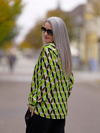 Emily Shirtbluse green sand: Frau mit grauem Haar trägt eine grün-schwarz gemusterte Shirtbluse mit kleinem Kragen, V-Ausschnittblende und 1/1 Ärmel. Sichtbare Lurex Tape-Details ergänzen den Retro-Print.