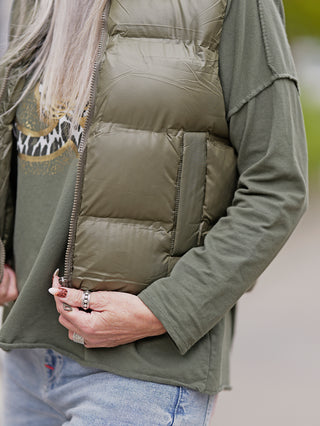 Nahaufnahme einer Frau, die die OFF#DLY West Ronja khaki Steppweste mit Stehkragen hält, zeigt die seitlichen Taschen und den Reißverschluss. Die Weste ist aus 100% Polyester gefertigt.