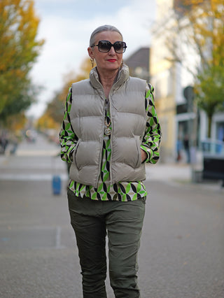 Frau trägt die OFF#DLY West Ronja taupe, eine kurze Steppweste mit Stehkragen und seitlichen Taschen. Sie kombiniert die Weste mit einer Sonnenbrille auf einem Gehweg.