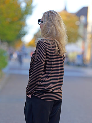 Frau trägt das OFF#DLY Ringelshirt in Schwarz/Braun, mit langen Ärmeln und geschnittenen Kanten, während sie auf dem Bürgersteig steht. Aus Viskose, mit Händen in den Taschen.
