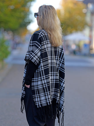 Eine Frau trägt den OFF#DLY Poncho Dalton in Schwarz/Weiß, ein stilvolles, kariertes Cape, perfekt für die Straße. Ideal für modische Outdoor-Looks und lässige Anlässe.