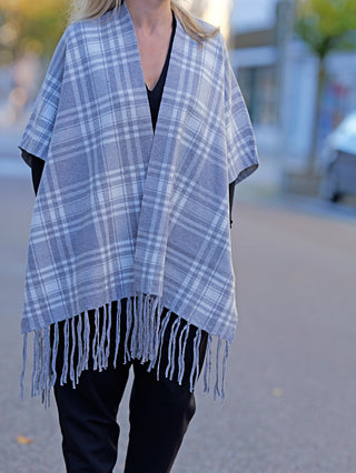 Eine Person trägt den OFF#DLY Poncho Dalton grau, der ein kariertes Muster zeigt. Der Poncho hat Fransen und verleiht dem Straßenmode-Look einen lässigen, stilvollen Touch.