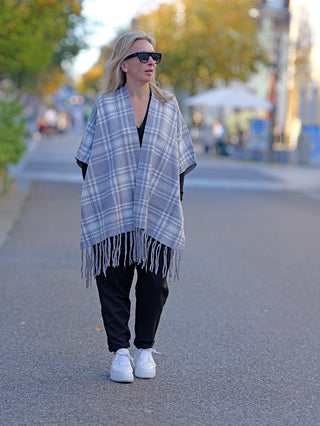 Frau trägt OFF#DLY Poncho Dalton grau, kombiniert mit weißen Sneakers, während sie auf dem Bürgersteig läuft. Der Poncho zeigt ein kariertes Muster mit Fransen.